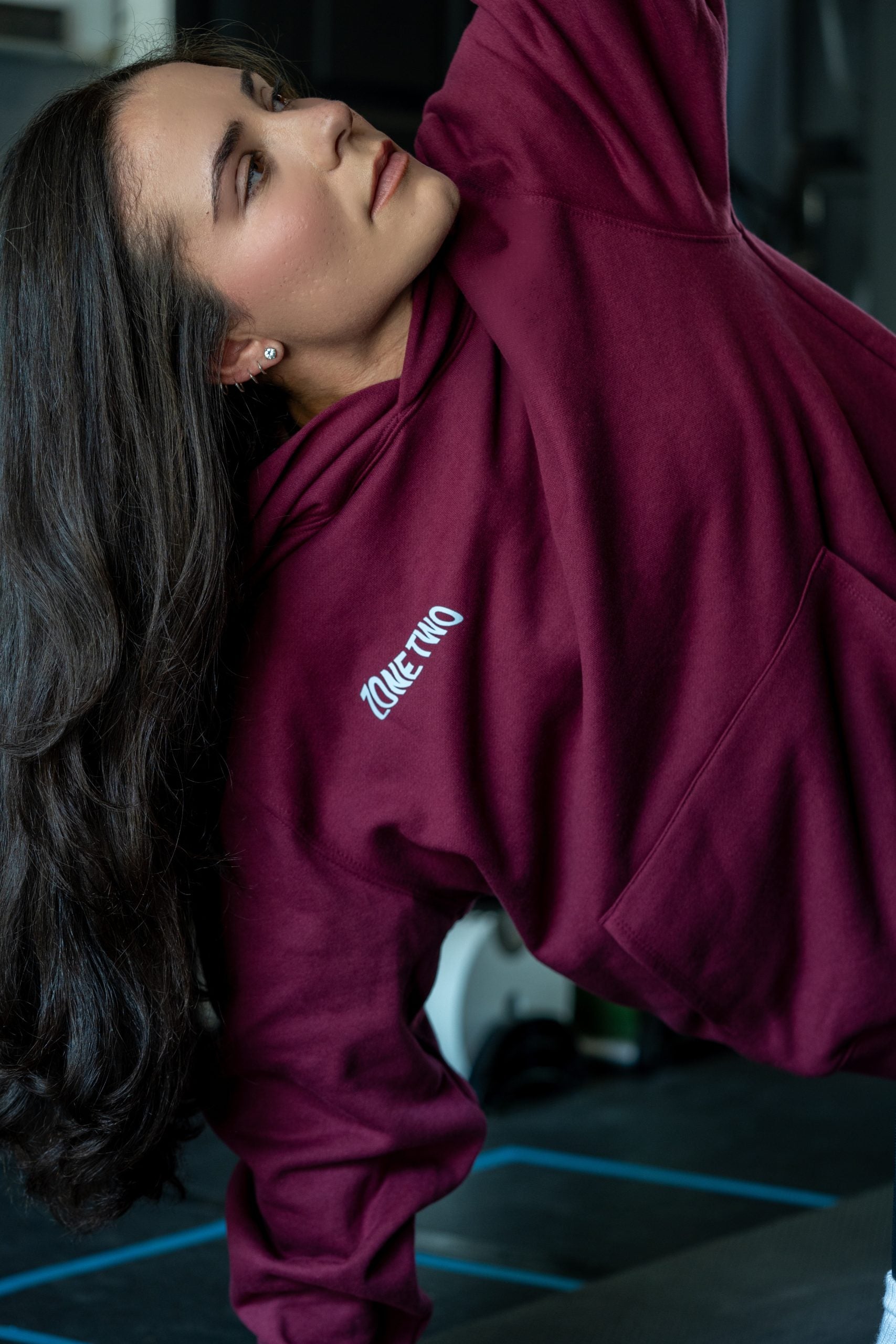 A woman in the burgundy Forward Hoodie in low light, with ZONE TWO printed at the sleeve.