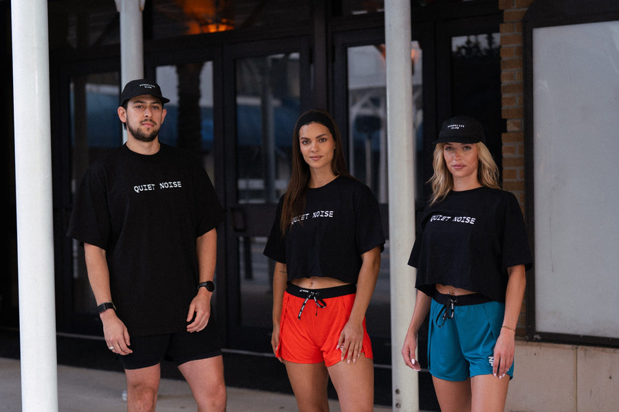 Three people outside a building in black Quiet Noise tees and caps.