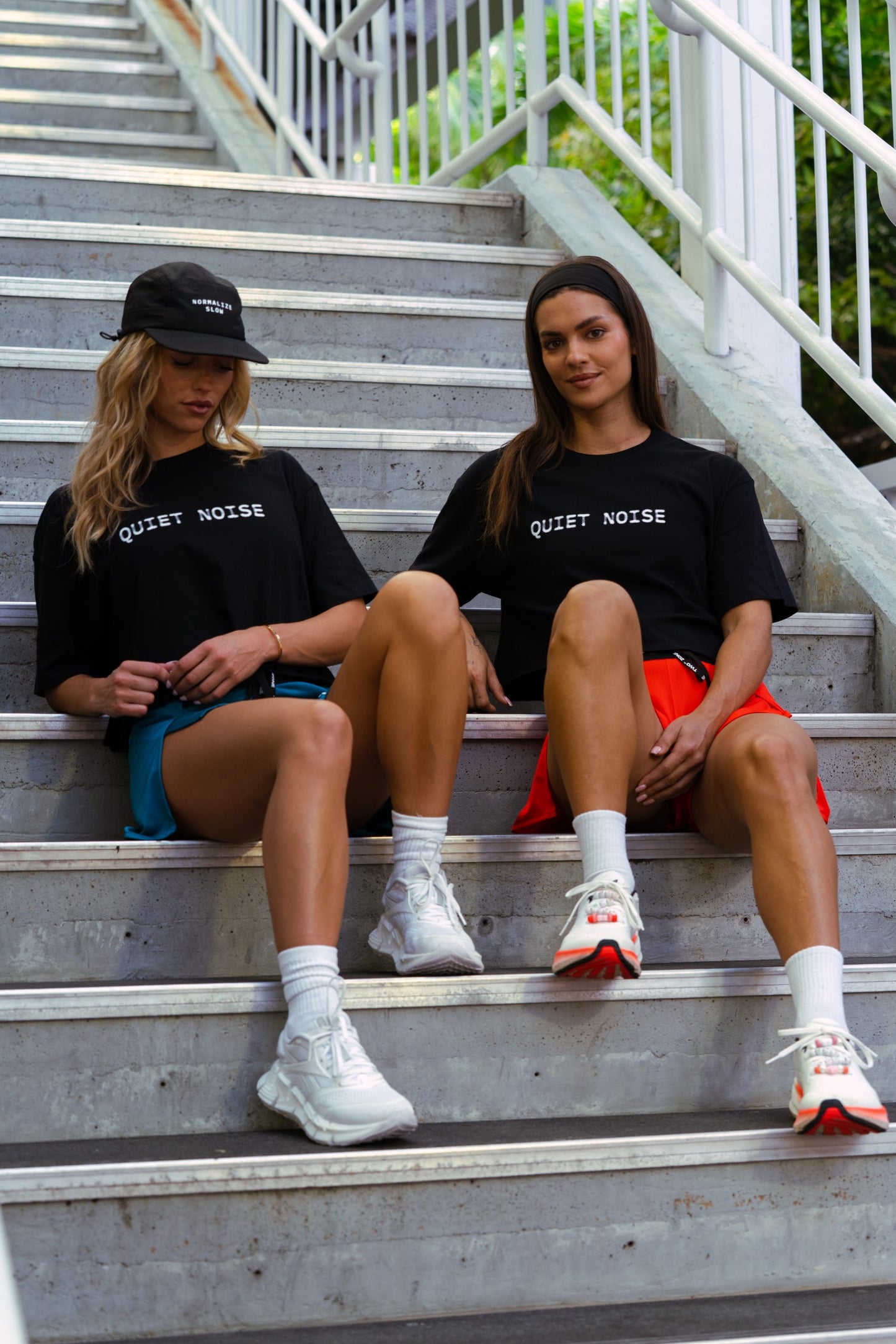 Two women seated on steps in black Quiet Noise Crop tops and caps.