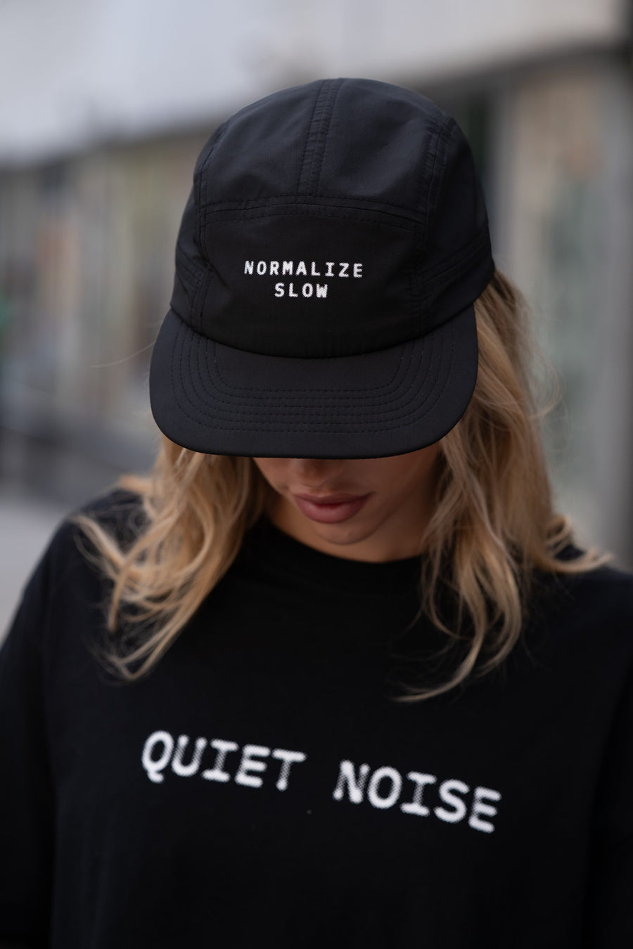 A woman in the black Normalize Slow Cap, printed NORMALIZE SLOW across the front panel, over a black QUIET NOISE top.
