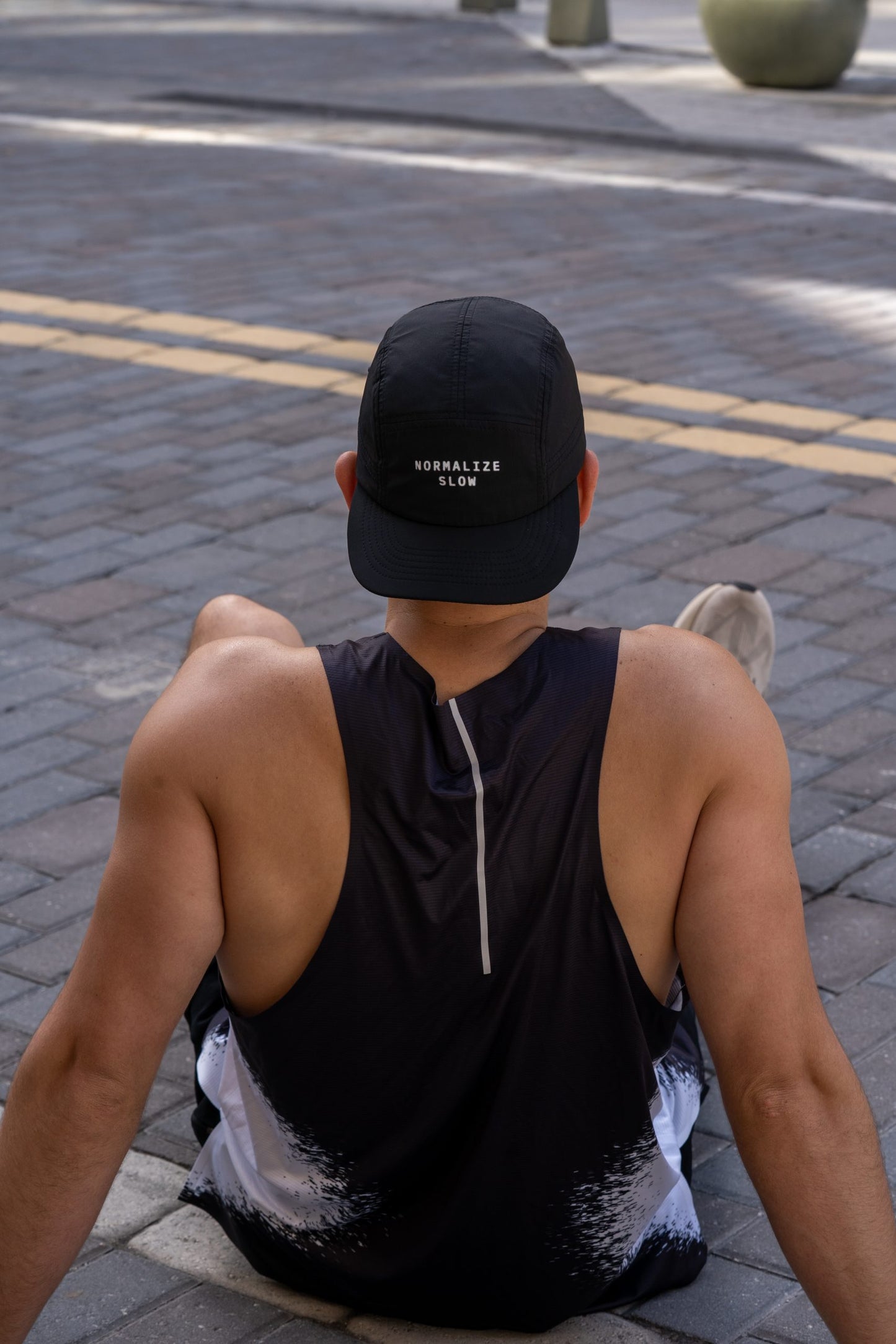 A man seated on a kerb seen from behind, wearing the Static Tank and a cap.