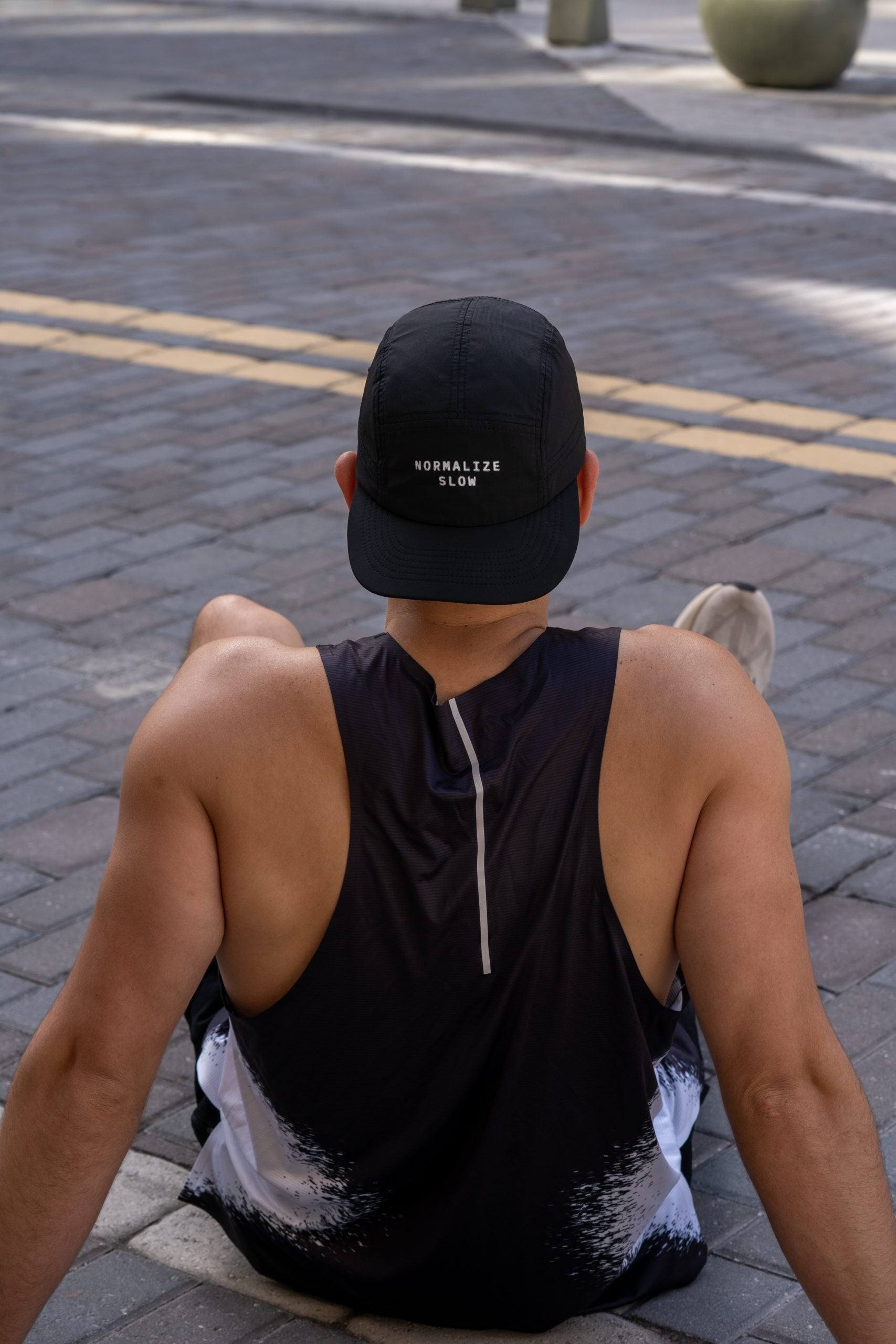 A man seated on a kerb seen from behind, wearing the Static Tank and a cap.
