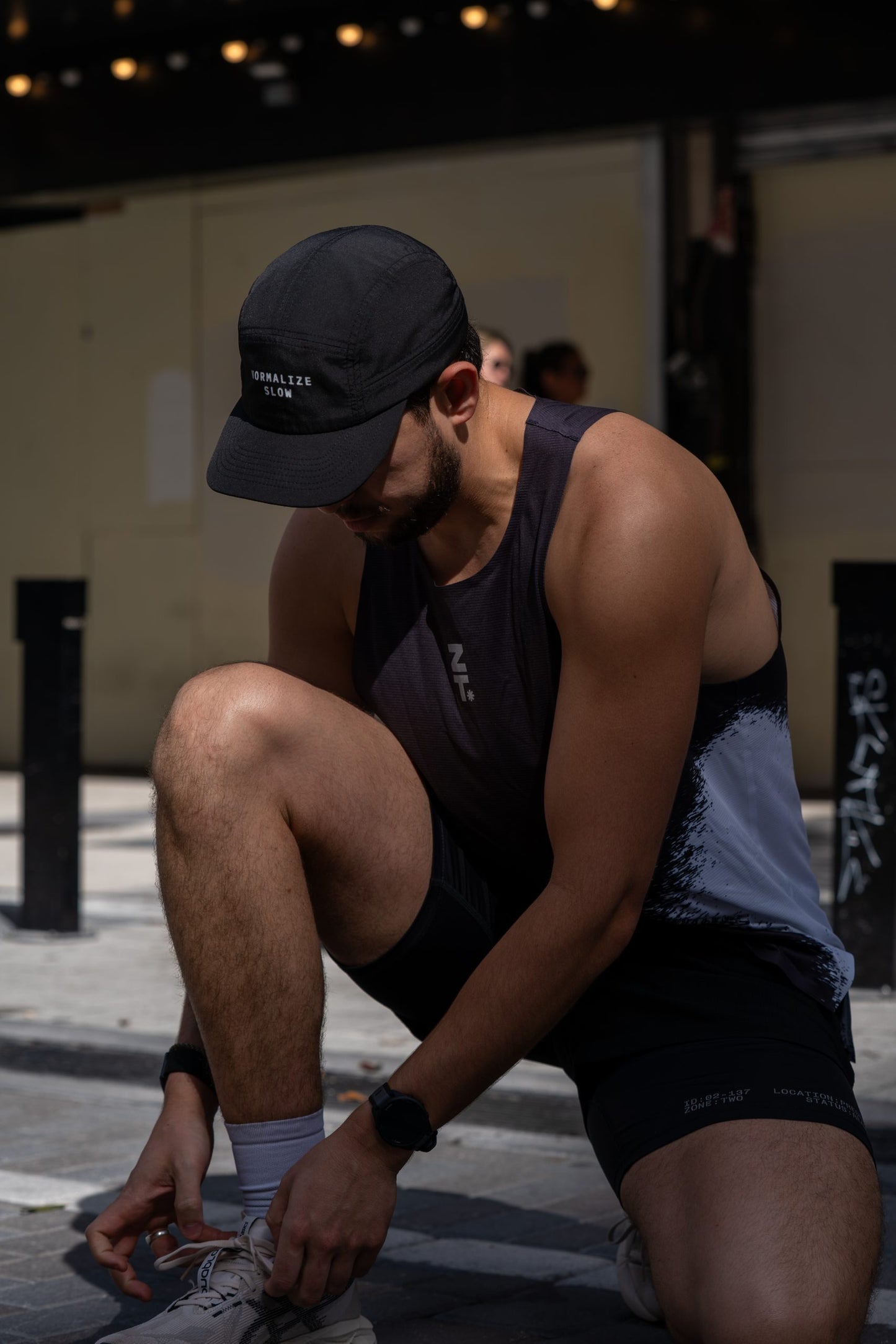 A man in the black Normalize Slow Cap crouching to tie a shoe on a street.