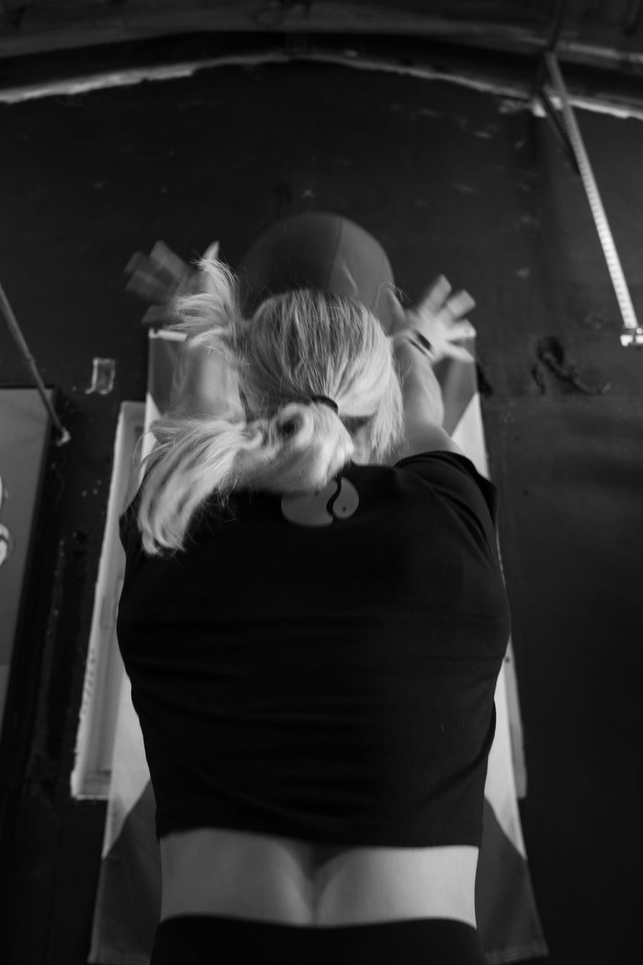 A black-and-white photograph of a person from behind with arms raised, wearing a dark cropped top.