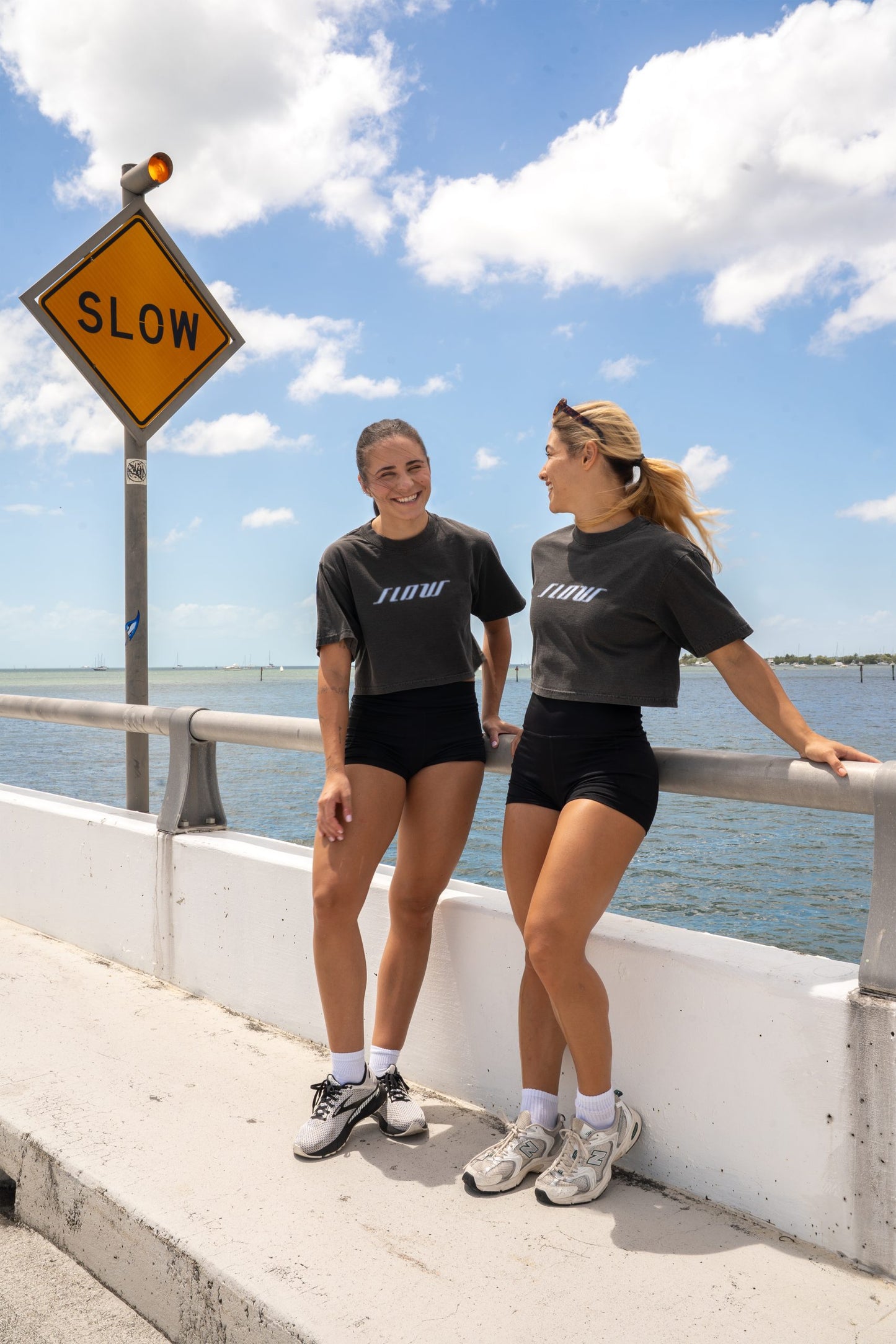 Two women in Slow Crop tops beside a road sign reading SLOW, at the waterfront.