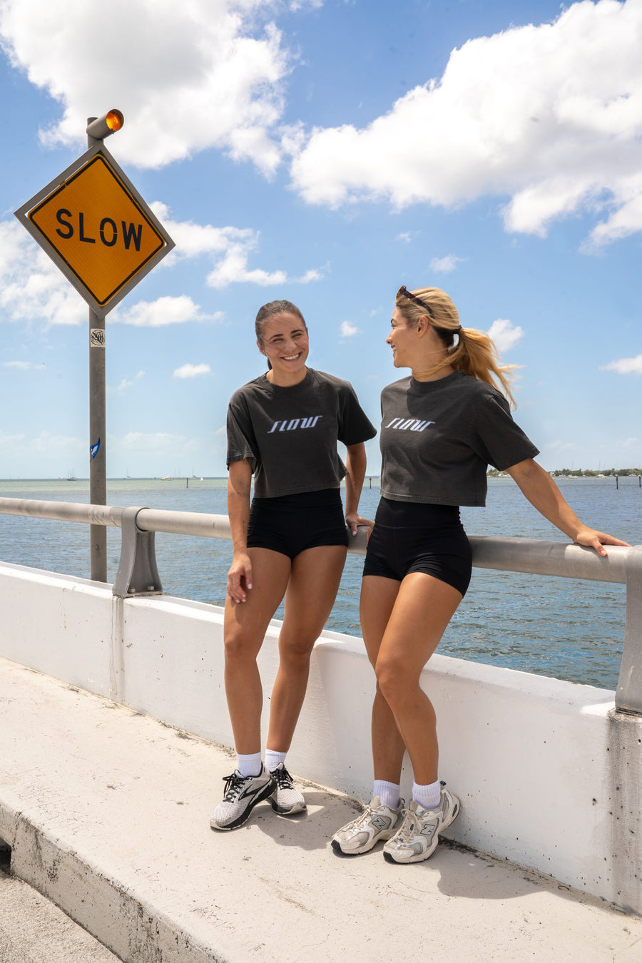 Two women in Slow Crop tops beside a road sign reading SLOW, at the waterfront.