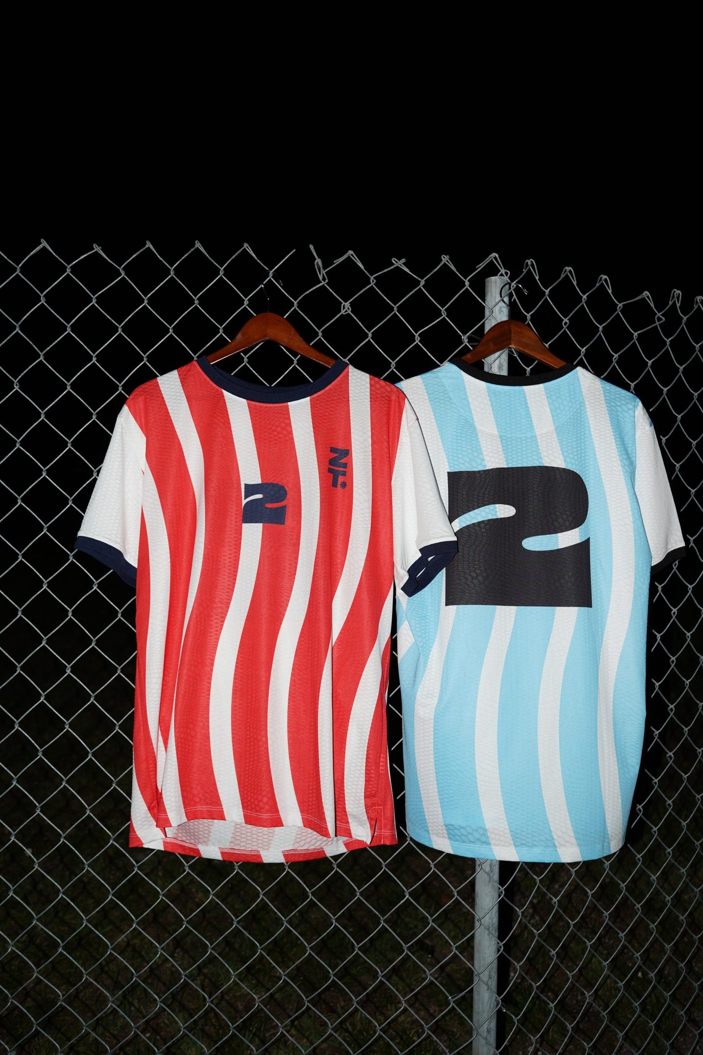 Both Nations Tee colourways hanging on a chain-link fence.