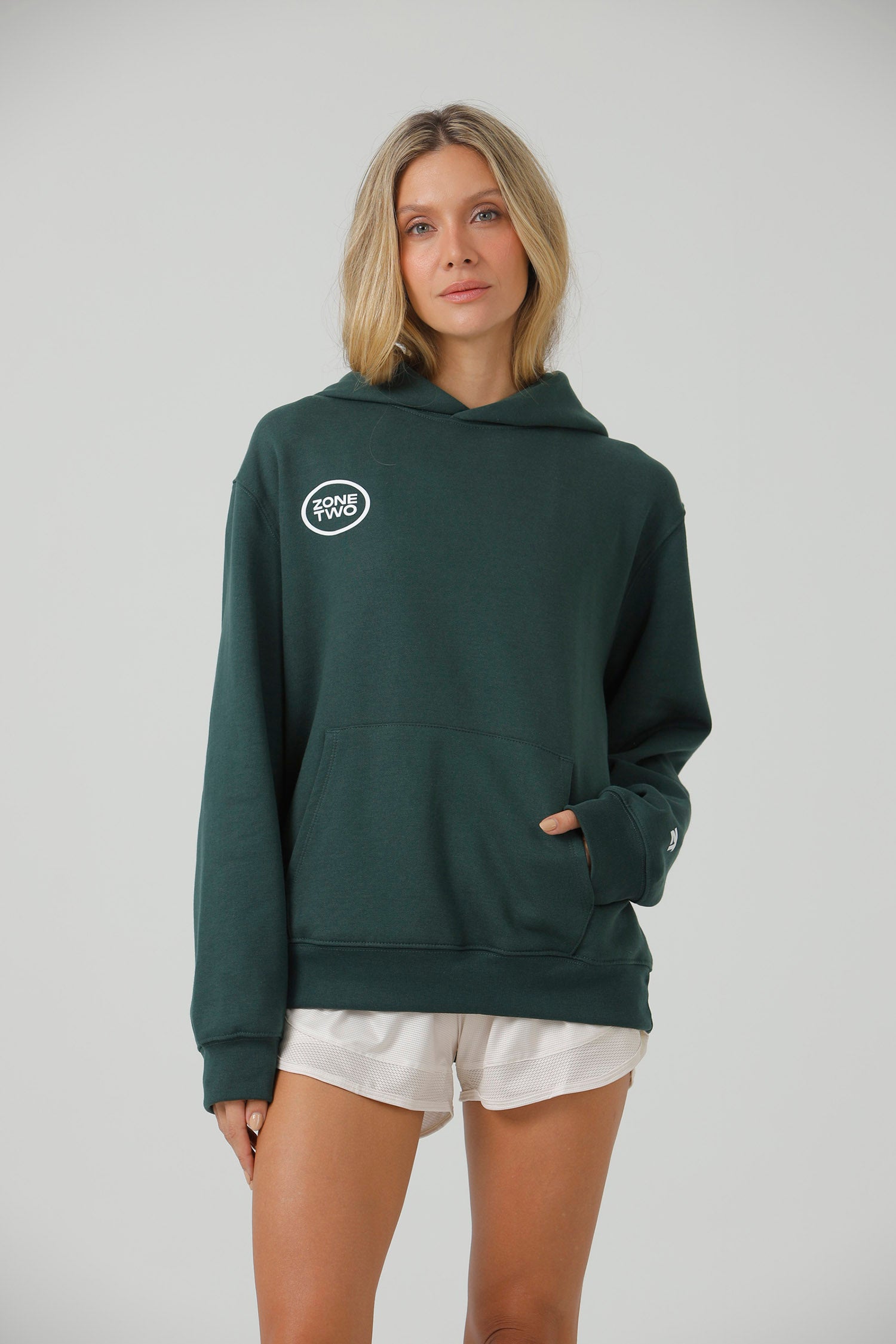 A model in the green Quiet Hoodie with hands in the front pocket, showing the oval mark at the chest.