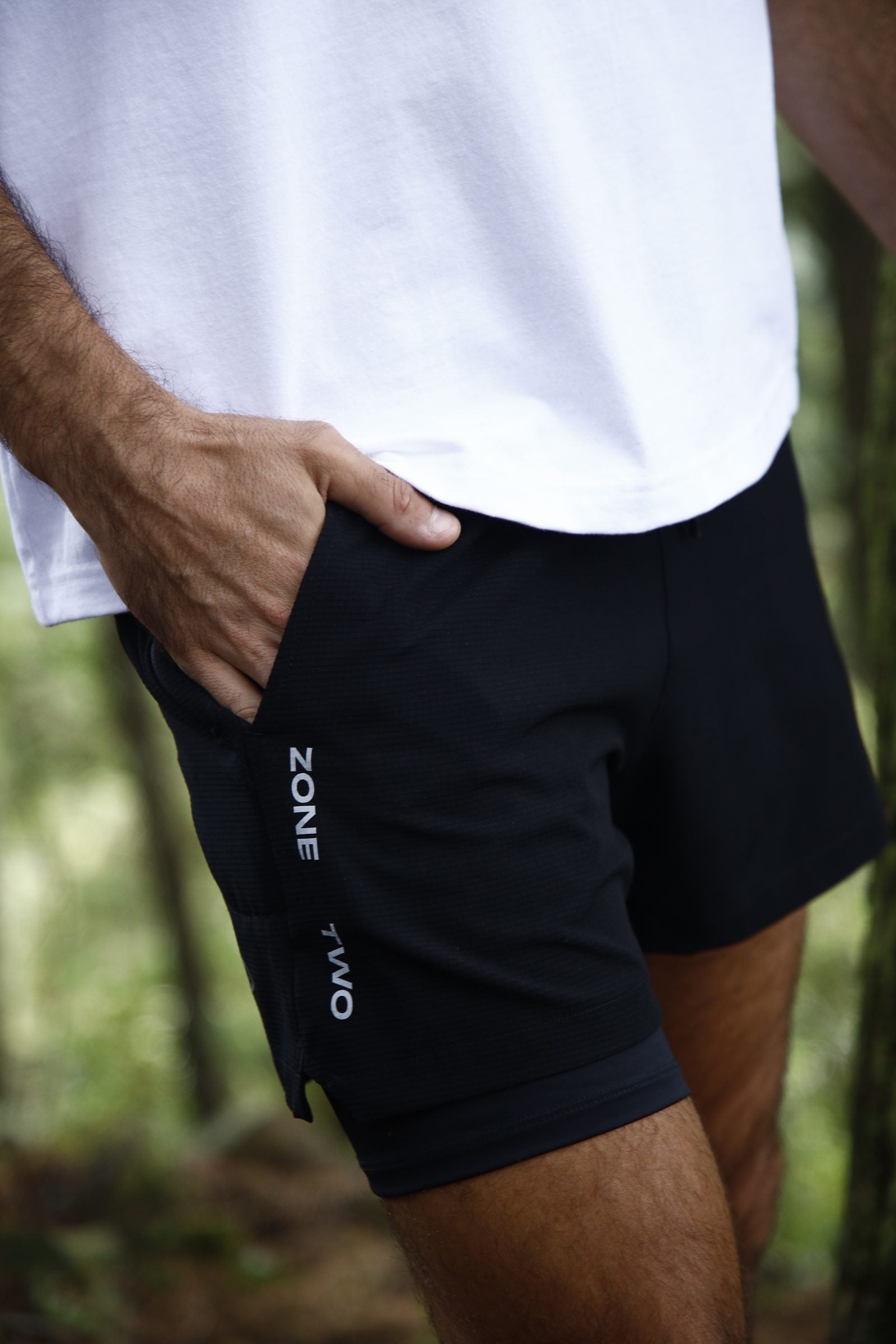 A man in the black Drift Shorts outdoors, hand at the hip pocket, Zone Two wordmark at the leg.
