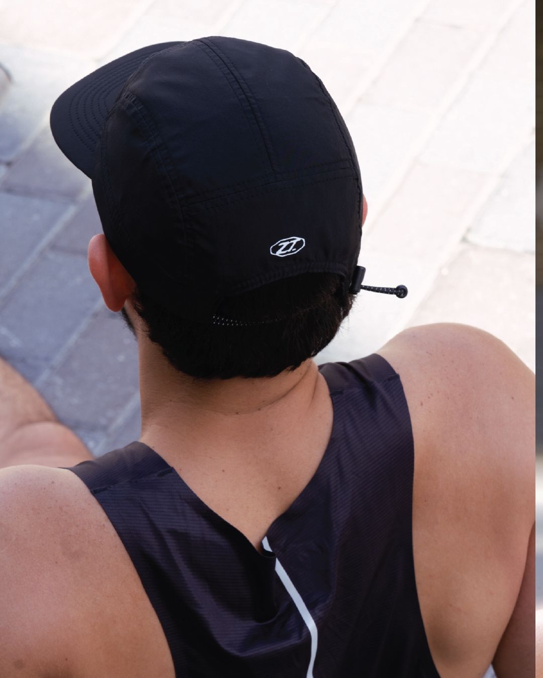 A man seen from behind wearing the dark Normalize Slow Cap and a sleeveless top.